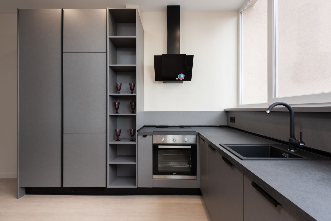 Interior of light gray kitchen corner furnished with cupboards and stone counter with built in oven and sink placed with big windows in apartment